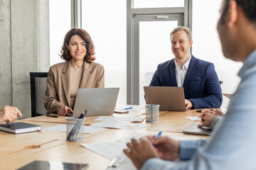 Three business professionals are sitting around a table in a modern office, using laptops and working on projects. Papers and writing utensils are spread out on the table