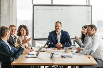 A group of business professionals sit around a large wooden conference table, engaged in a meeting....
