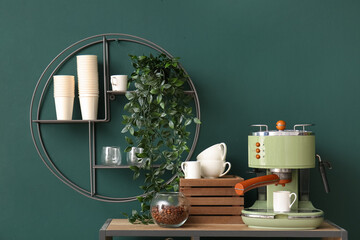 Modern coffee machine, cups and jar with beans on table near green wall