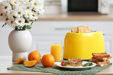 Modern toaster with crispy bread slices, glass of juice, orange and delicious sandwiches on table in kitchen