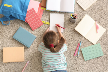 Cute little girl with backpack, notebooks and pencil lying on floor and studying at home