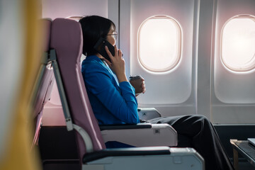 Professional woman in business attire conversing on mobile phone and holding coffee cup while traveling on an airplane.