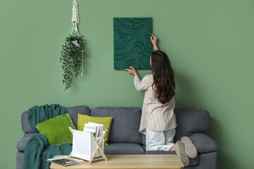Young pretty woman hanging picture on green wall at home