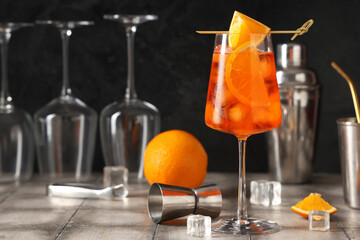 Glass of Aperol spritz cocktail with ice cubes and shaker on grey tile table