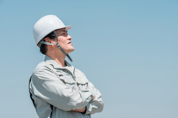 青空の下でヘルメットを被って空を見上げる作業着姿のアジア人男性作業員
