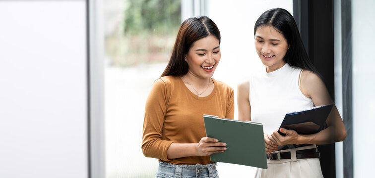 Two happy asain business women talking and discussing, standing in creative green office space.