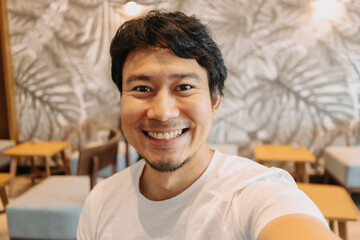Happy asian man with beard smiling and taking selfie while sitting in cafe.