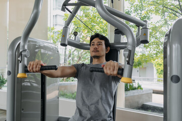 Fitness asian Thai man sitting, focused using chest and shoulder press machine at gym, building...