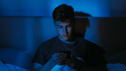 Young man using a phone in dark room with blue light symbolizing insomnia or screen addiction.