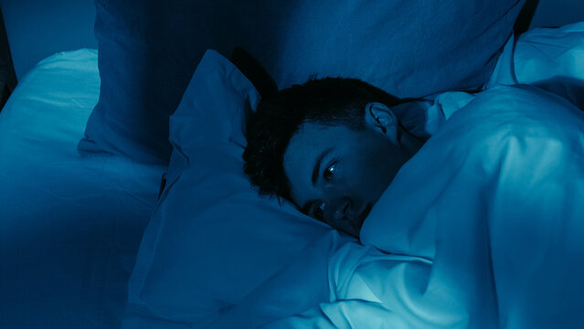 Exhausted man experiencing loneliness insomnia lying awake in bed under dim blue lighting.