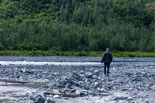 A man in a black jacket walks alone along a rocky riverbed surrounded by dense forest in the Kenai Peninsula wilderness.