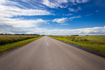 A long, straight road leads through vibrant fields, surrounded by a brilliant blue sky adorned with fluffy clouds on a sunny day.