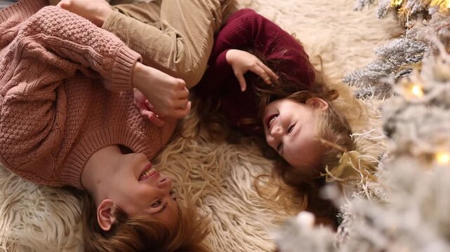 A young mother and daughter 5-6 years old in casual clothes lie on a soft carpet in a cozy, and decorated Christmas living room at home against the backdrop of a Christmas tree.