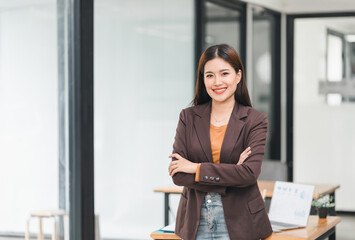 Confident asian businesswoman stands with arms crossed in modern office, wearing blazer and smiling...
