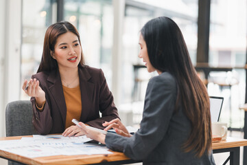 Two businesswomen in formal attire having discussion at desk with charts and graphs, focusing on data analysis and teamwork.