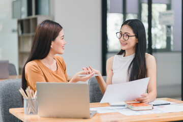 Two businesswomen converse in a contemporary office, sharing ideas and documents while seated at working desk.