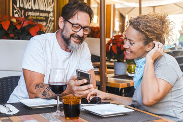 Modern adult couple sitting at table restaurant watching smartphone to leave a review feedback after lunch. Choosing the best pizzeria before eat reading comments on line on website on the phone