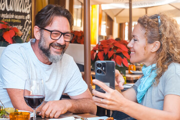 One couple watching phone display sitting at the restaurant before lunch. Reading or writing review online on web page. Calling friends to invite to have lunch together. Dating man and woman happy