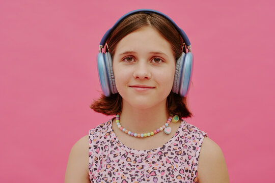 Medium closeup of modern gen alpha teen girl wearing headphones standing against pink wall background and looking at camera