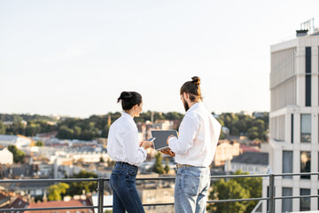 Business colleagues standing on rooftop, discussing project using technology devices. Urban cityscape view in background provides professional setting. Teamwork and collaboration concepts represented.