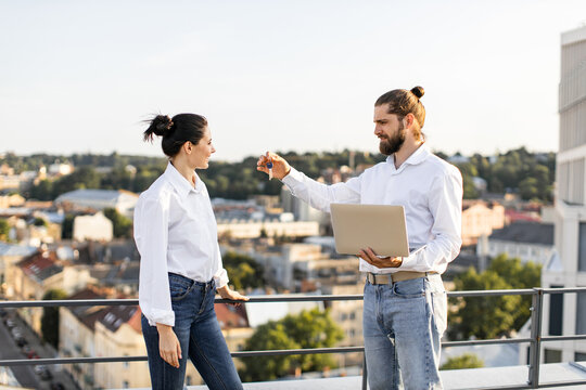 Two people standing on urban rooftop exchanging house keys. Man holds laptop while handing keys to woman, representing new homeownership, real estate, and modern lifestyle.