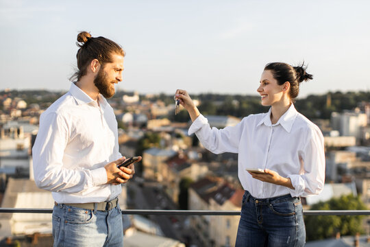 Real estate agent handing keys to new homeowner on rooftop with cityscape background. Man and woman in casual attire smiling, holding electronic devices, and celebrating new property transaction. - Powered by Adobe