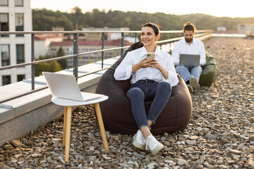 Two people working remotely on rooftop terrace using laptops and smartphones. Outdoor workspace promoting relaxation and productivity, surrounded by urban landscape.