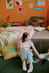 Obraz premium Vertical shot of teenager with disability sitting on floor in her cozy bedroom and studying via laptop, copy space