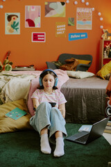 Obraz premium Vertical shot of teen girl with disability in headphones choosing playlist on smartphone while sitting on floor against her bed and doing homework