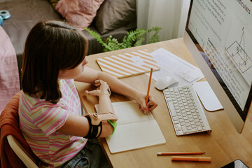 High angle view of teenager with prosthetic arm sitting at desk in her room and writing in copybook during online class