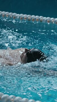 Vertical tracking from above slowmo shot of Caucasian athletic man swimming backstroke in pool water