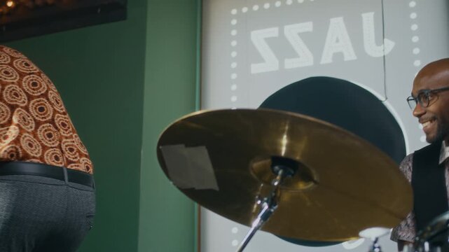 African American man with conga drum entering music studio, greeting his bandmate with handshake and hug, then taking his seat ready to start rehearsal