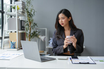 Asian businesswoman sitting, relaxing, holding a hot coffee mug, working with laptop on table, online finance, documents, taxes, reports, marketing, statistics, business analysis research concept.