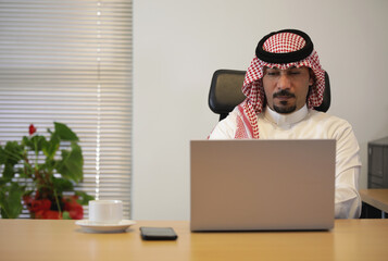 Arab man working on laptop in the office, watching stock market and talking on the phone and in...