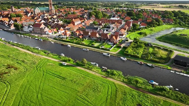 Camera pans along the riverside of Ribe with docked boats, tilting up to reveal the historic town Ribe with its majestic cathedral and charming red-roofed buildings.