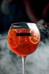 A refreshing Aperol Spritz cocktail in a wine glass with ice cubes and orange slices. The drink is set against a dark, moody background.