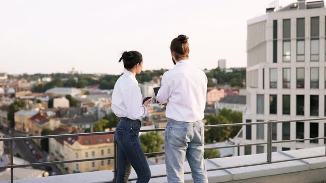 Two business colleagues discussing project on rooftop with city view. One points in distance while other holds laptop, emphasizing collaboration, innovation, and teamwork.