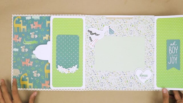 Opens baby themed scrapbook with unique design, flip page with central space for photograph, surrounded by kiddish designs.