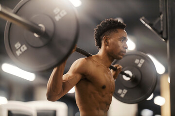 Strong shirtless muscular black bodybuilder with barbell on shoulders at gym.