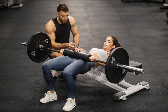 Muscular sportswoman doing hip thrusts on bench with barbell and her coach is motivating her.