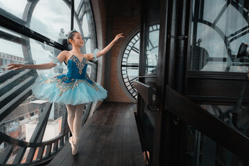 A young girl in a blue tutu is dancing in front of a clock. The clock is large and has a gold face. The girl is wearing a tiara and he is performing a ballet routine © phaitoon