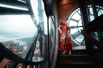 A woman in a red dress is playing a cello in front of a clock. The clock is large and has a unique design. The scene is set in a building with a view of the city