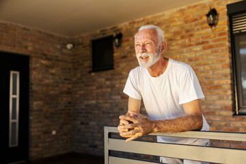 Senior man standing on porch and leaning on fence and enjoying view on his backyard.