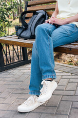 Young woman in casual clothes sitting on the bench in park. Girl in blue jeans and white shoes with backpack relaxing in nature. Tranquility in big city lifestyle. Person waiting alone on the bench.