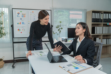 Two Asian businesswomen cheerfully discussing plans in office using calculator and laptop, standing in front of a whiteboard talking.