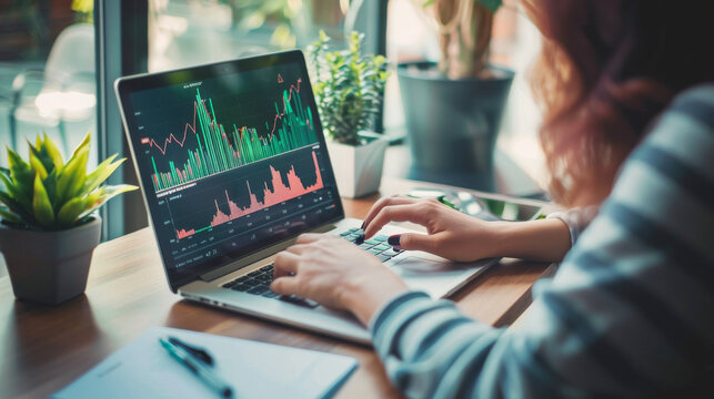 A person analyzes fluctuating stock market graphs on a laptop, surrounded by plants on a wooden desk in a bright, modern office.