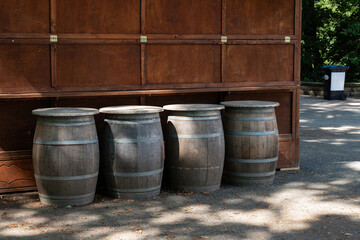 Four wooden barrels are lined up against a wooden wall in a park, basking in the daylight amidst scattered foliage.