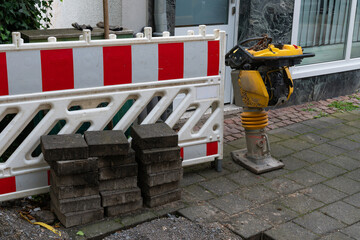 A construction site featuring a stack of paving blocks, a compacting machine, and a red and white...
