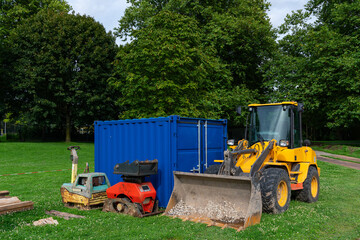 Obraz premium A yellow loader and red machine sit beside a blue storage container on green grass in a park under clear skies.
