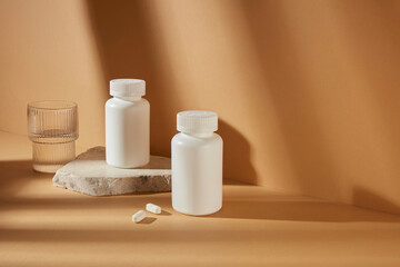 Medicine mockup for promotion, two white unbranded medicine bottle featured on brown background with a glass of water beside. Blank space for adding elements of designing, high angle shot